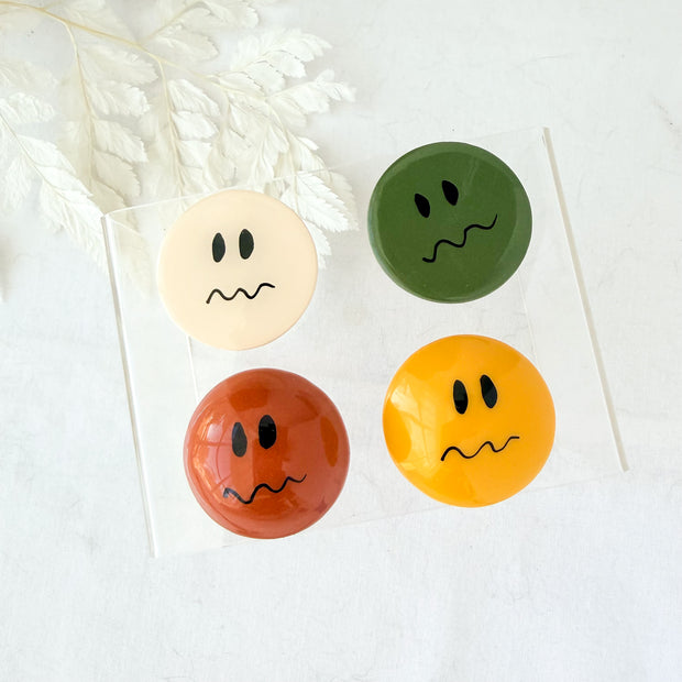 Round polymer clay fridge magnets with worried, wavy-mouth faces in cream, olive, terracotta, and mustard.